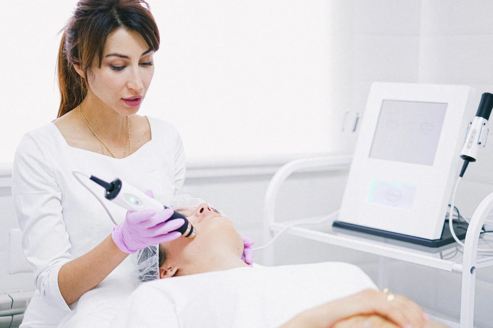 A clinician in a bright treatment room guiding a skin device across a patient's cheek